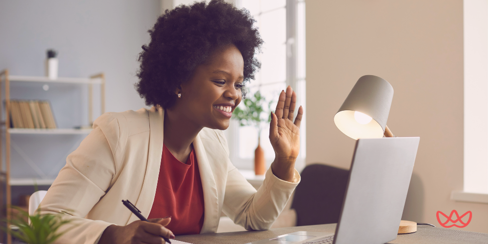 A businesswoman with a joyful expression waves at her laptop screen, engaged in a friendly virtual meeting from a well-lit office space.