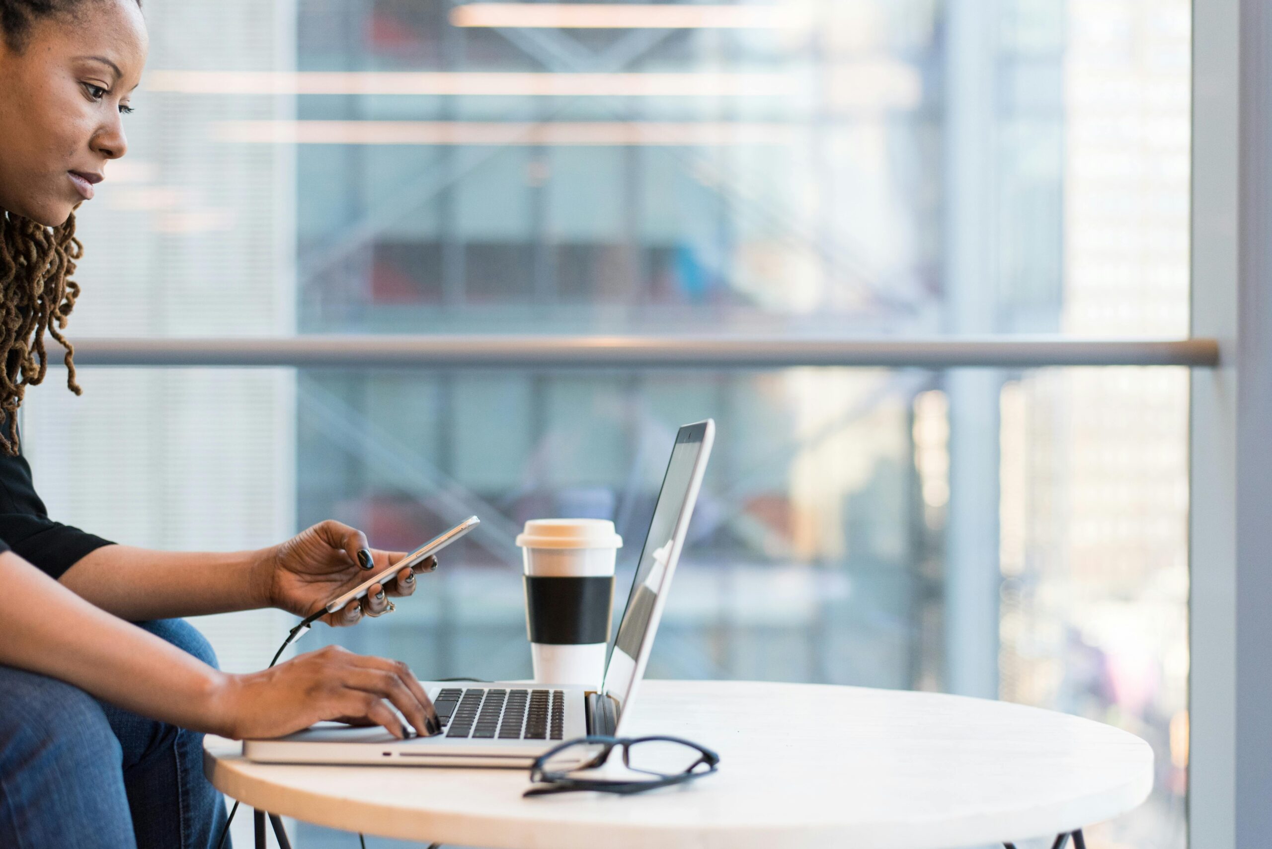 Administrative virtual assistants at work, with a focus on a person sitting at a table with a laptop, smartphone, and coffee cup, in an office environment with large windows.