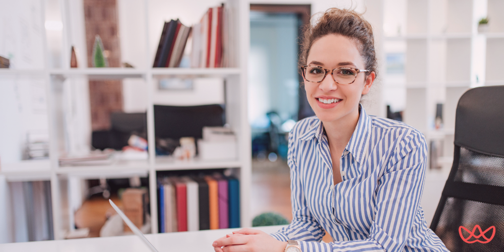 A smiling woman in glasses sitting at her office desk, preparing for a discussion on interview questions for office admin positions.