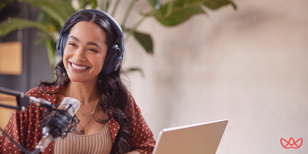 A smiling woman wearing headphones and speaking into a microphone while using a laptop, supported by a podcast virtual assistant.