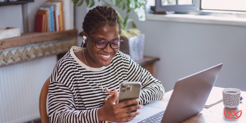 A smiling professional woman wearing glasses and earphones, sitting at her desk with a laptop and a phone, embodying the benefits of why hire a virtual assistant.