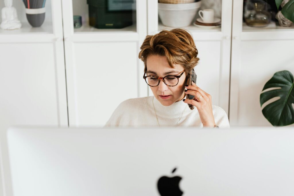 A woman speaking on the phone while working on a desktop computer in a modern home office, representing the multitasking nature of a virtual business manager.