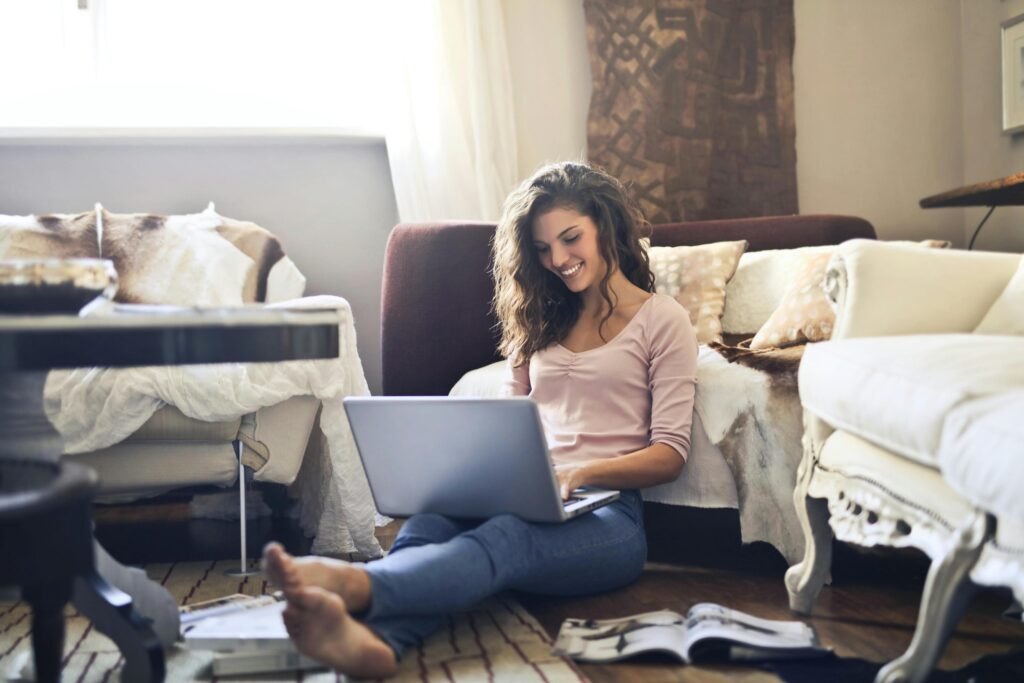 A smiling woman sitting on the floor with a laptop, managing tasks for a content creator assistant role in a cozy home environment.