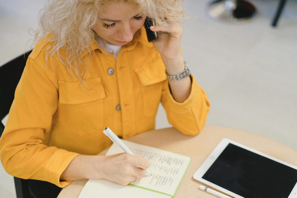 A woman in a yellow shirt taking notes during a phone call, reflecting the multitasking capabilities of real estate virtual assistance.