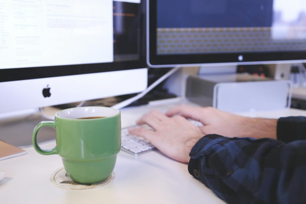 Close-up of hands typing on a keyboard with dual monitors and a coffee cup, symbolizing a short-term rental virtual assistant managing tasks remotely.
