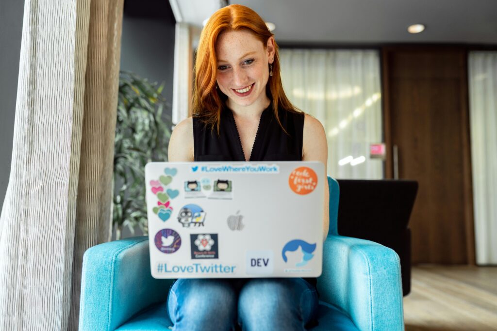 A red-haired woman smiling while working on a laptop covered with stickers, representing the concept of "why do companies engage in outsourcing."
