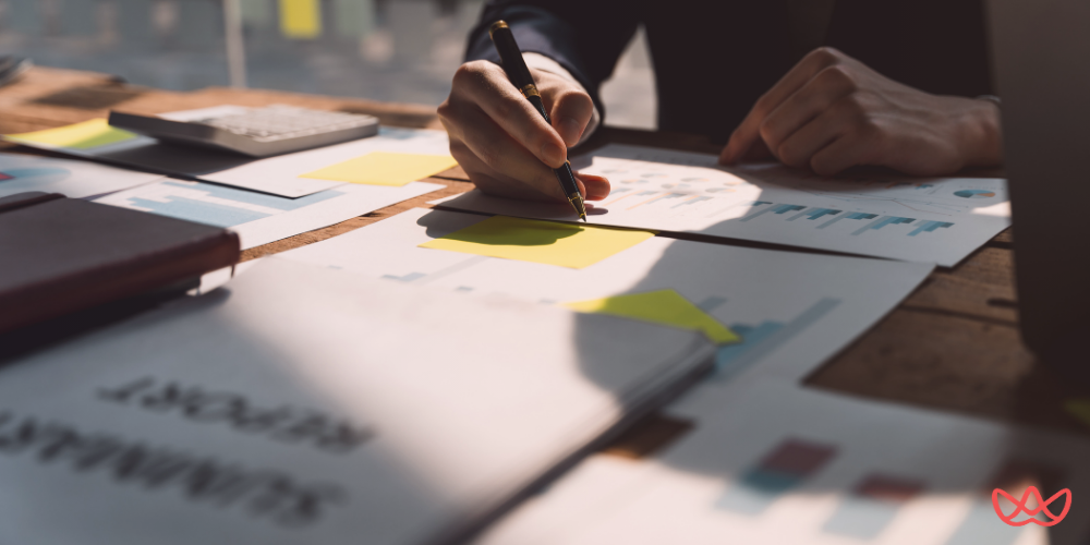 An account manager key skills summary is being reviewed on a desk cluttered with documents, charts, and a calculator. A person is writing notes on a yellow sticky note while analyzing various reports and graphs.