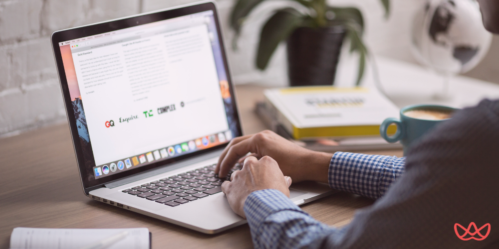A person in a checkered shirt working on a laptop at a wooden desk, viewing a webpage with marketing-related content. The setup includes books, a coffee cup, and a plant, representing the workspace of an Email Marketing Copywriter Key Skills Summary.