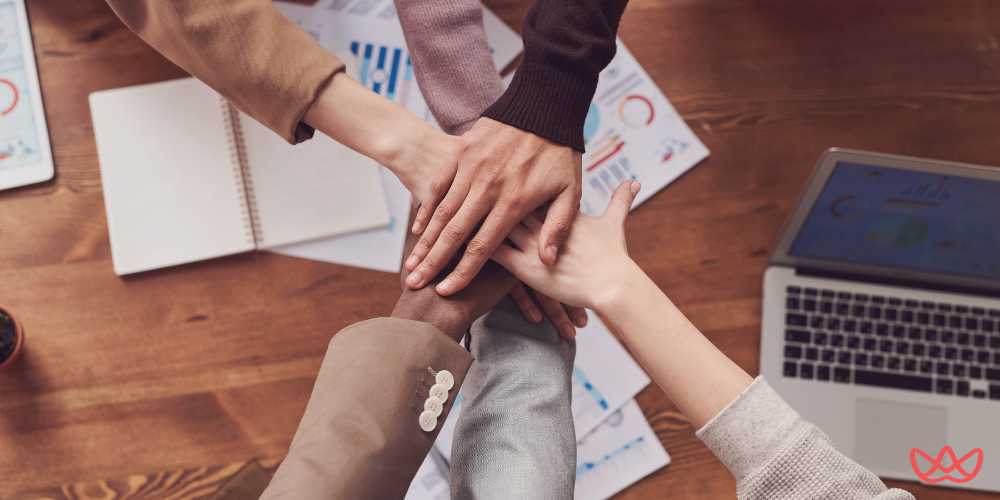 A group of diverse hands coming together over a wooden table with business documents, a notebook, and a laptop, symbolizing teamwork and collaboration.