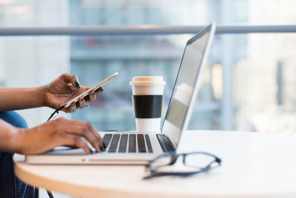 A person working on a laptop while holding a smartphone, with a coffee cup and glasses on the table, illustrating the multitasking nature of a Display Advertising Manager Key Skills Summary.