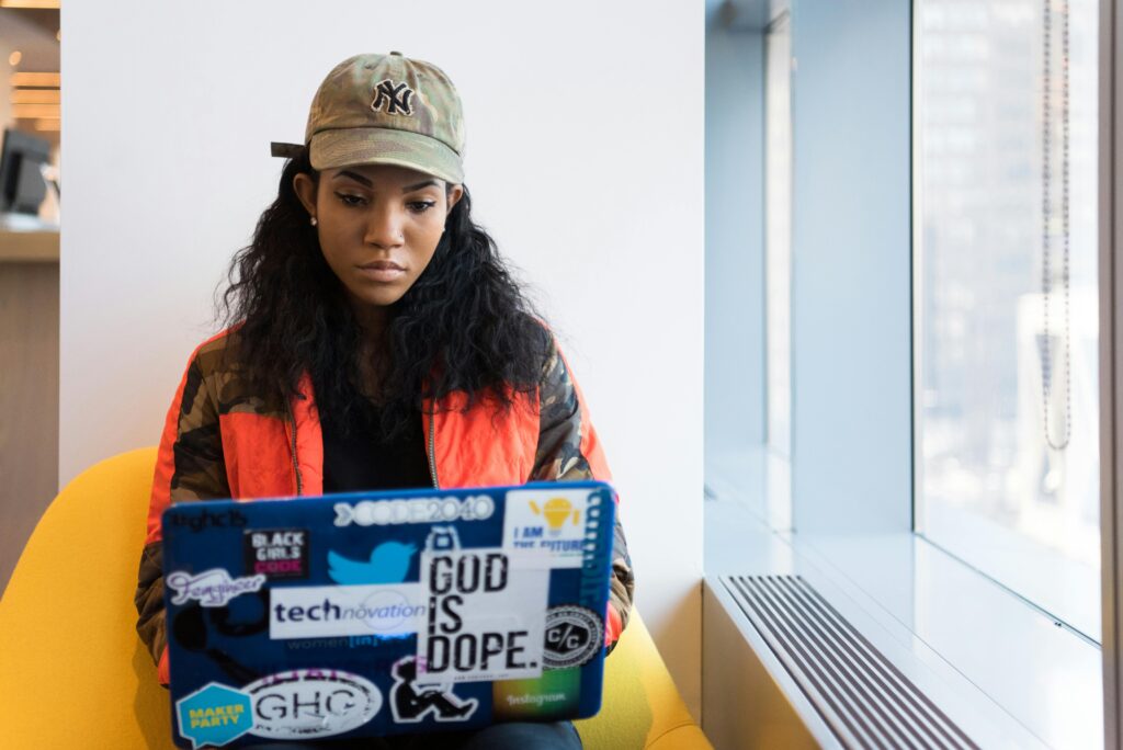 A woman in a NY cap and orange jacket working on a laptop decorated with tech stickers, representing backend developer key skills summary in a modern office setting