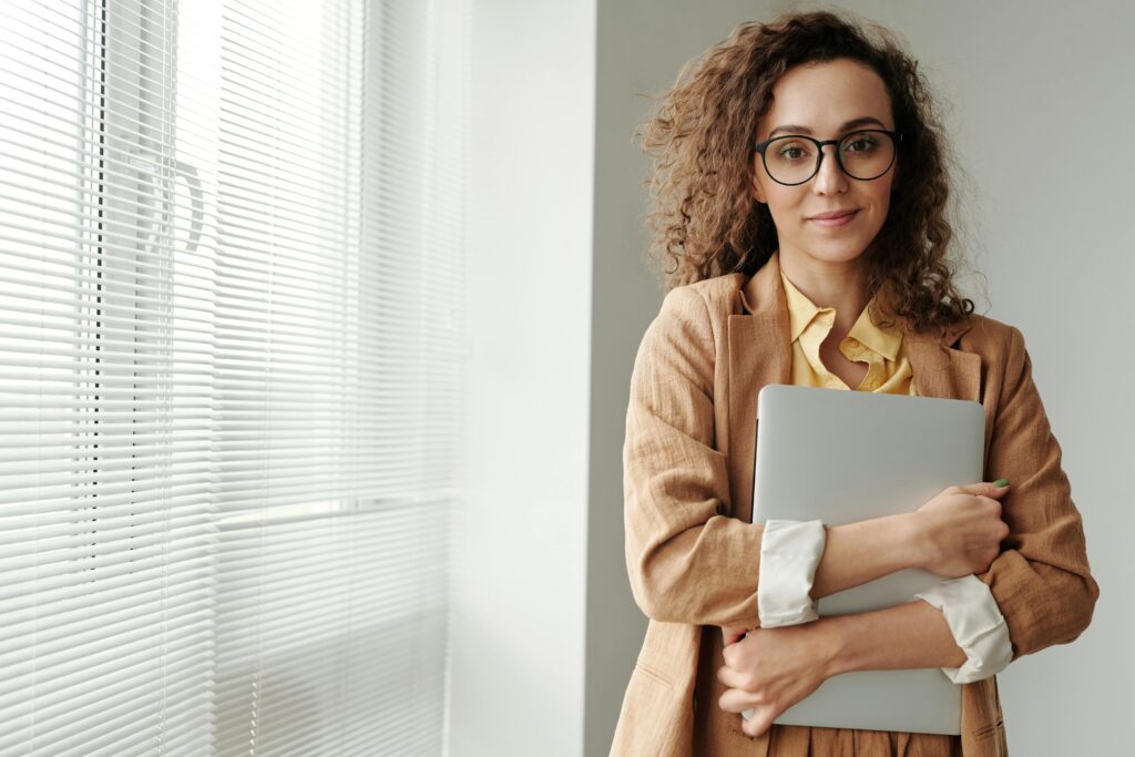 Professional woman in business attire holding a laptop, wearing glasses and a tan blazer, embodying business development manager key skills summary with confident demeanor