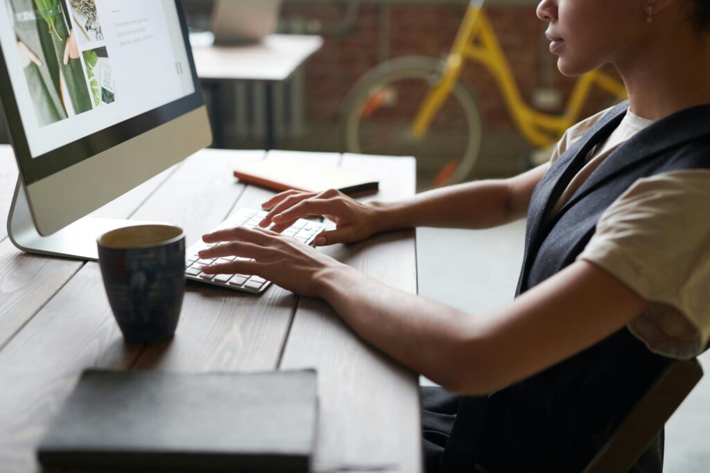 A digital product designer working on a desktop computer, typing on a keyboard with a coffee mug and a notebook on the wooden desk.