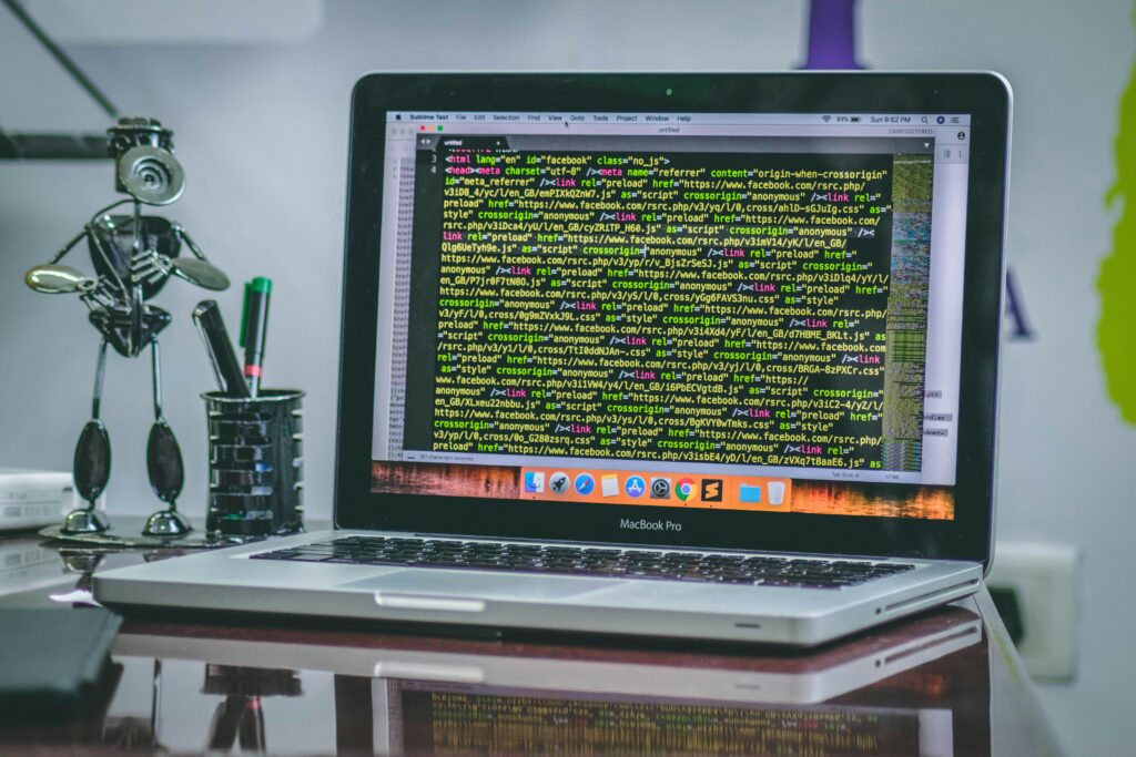A MacBook Pro displaying HTML code on the screen, surrounded by a robot figurine and a pen holder, representing the workspace of a frontend developer key skills summary.