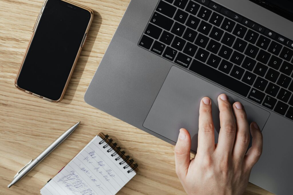 A client success manager key skills summary is being prepared on a laptop, with a smartphone, pen, and notepad nearby on a wooden desk.
