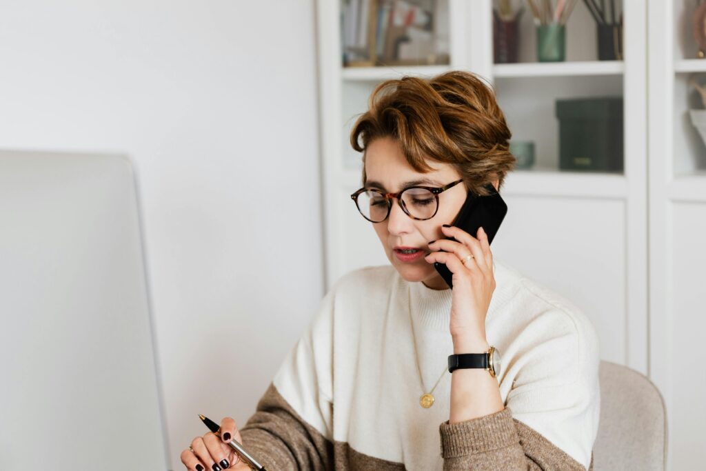 A marketing coordinator key skills summary: A person is sitting at a desk, holding a phone to the ear with one hand and a pen in the other, appearing to be engaged in a work-related task. The person is wearing a white and brown sweater and a wristwatch, with a blurred face for privacy.