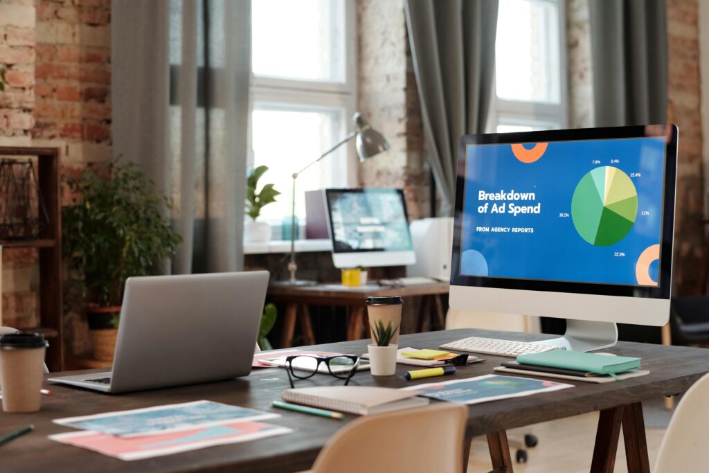 A modern office workspace with a computer screen displaying a pie chart titled "Breakdown of Ad Spend" from agency reports, highlighting the Display Advertising Manager Key Skills Summary.