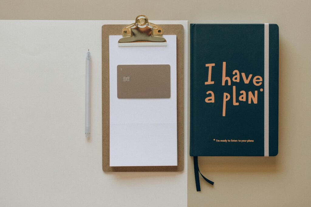 A workspace setup featuring an account manager key skills summary. The image includes a white pen, a clipboard with a blank sheet of paper and a credit card, and a dark green notebook with the text "I have a plan" on the cover.