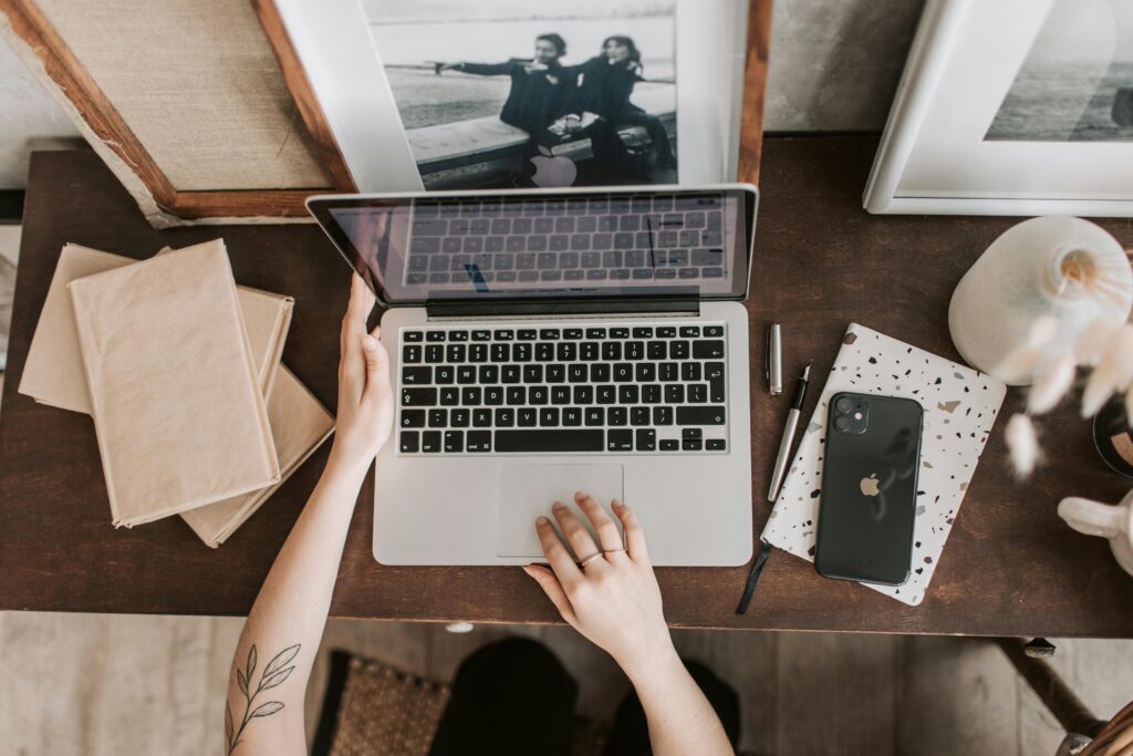 Overhead view of a person working on a laptop at a wooden desk with a notebook, smartphone, and framed photos.