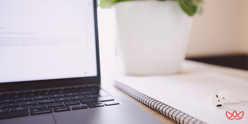 A modern workspace featuring a laptop with an open document, a spiral notebook, and wireless earbuds on a desk, with a potted plant in the background. This setup emphasizes a productive environment for programmatic marketing activities.