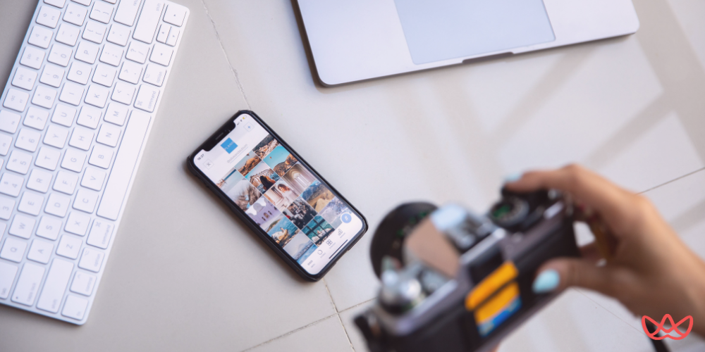 Social media manager capturing content with a camera, surrounded by a workspace including a smartphone, laptop, and keyboard.