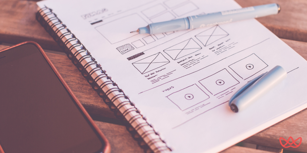 UX UI designer planning a website layout with detailed wireframe sketches in a notebook, alongside a smartphone and pen on a wooden desk.
