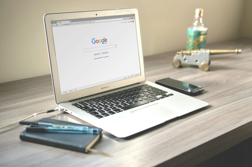 A MacBook Air displaying the Google homepage rests on a wooden desk beside a smartphone, a notebook with pens, and a small decorative cannon, reflecting an SEO content writer key skills summary.