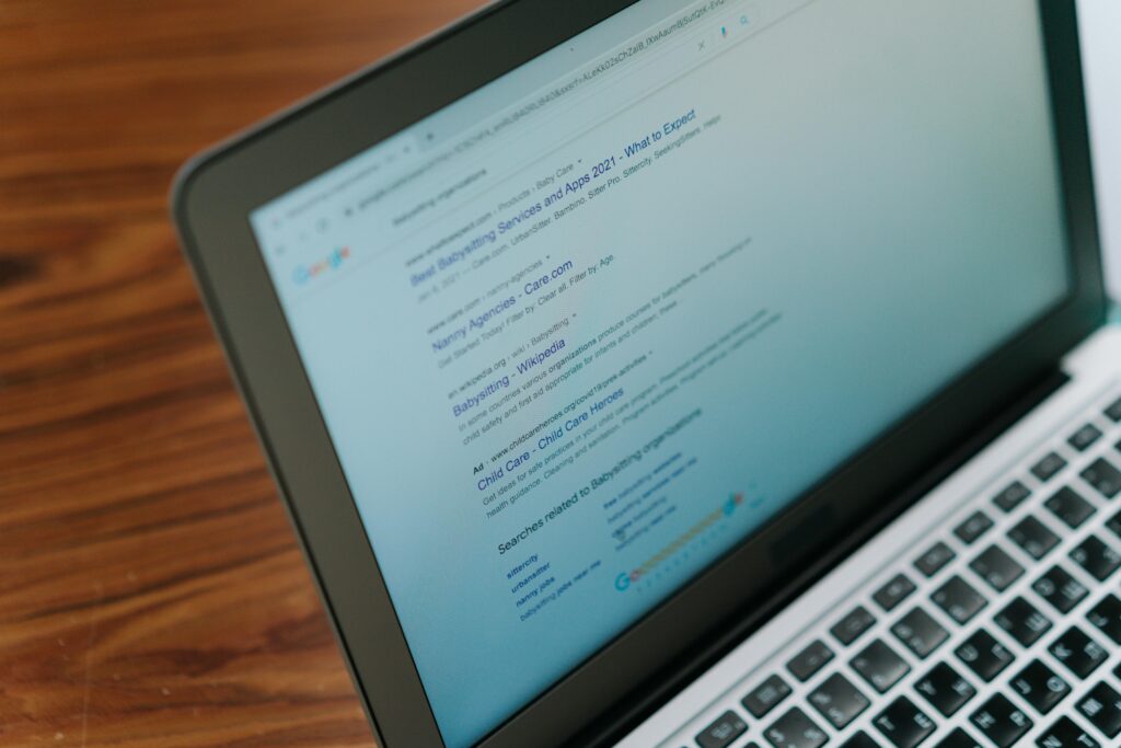 Laptop screen displaying Google search results with blurred search terms related to babysitting and child care services, viewed from an angle on a wooden surface.