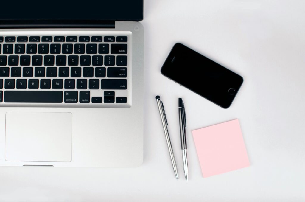 Minimalist workspace with laptop, smartphone, two metallic pens, and a pink sticky note, representing a Content Strategist's organized digital workflow.