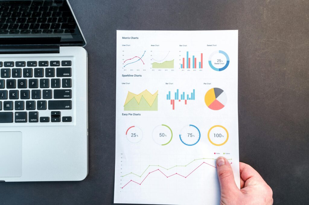 A laptop keyboard next to a hand holding a sheet of paper filled with various colorful charts and graphs, illustrating an SEO analyst key skills summary.