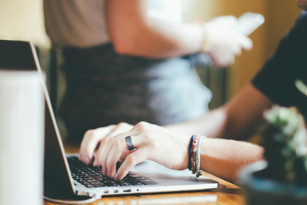 SEM director key skills summary: Hands typing on a laptop keyboard with a person in the background holding a smartphone.
