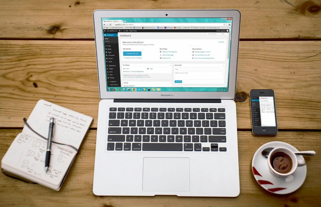 Workspace of a WordPress developer key skills summary with a MacBook Air showing the WordPress dashboard, a notebook with handwritten notes, a pen, a smartphone displaying the WordPress app, and a coffee cup on a wooden table.