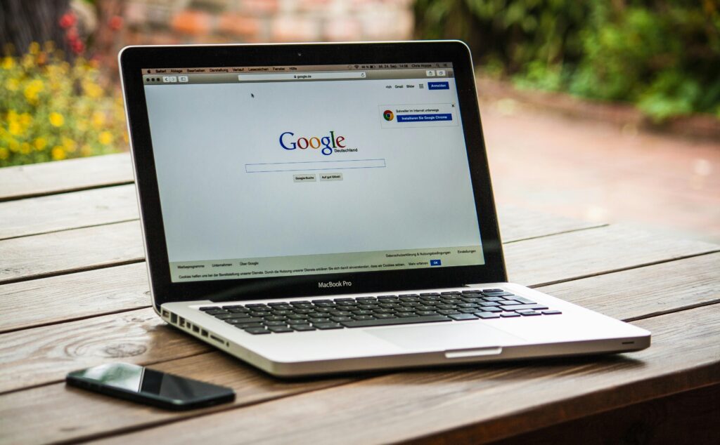 A MacBook Pro displaying the Google homepage with "google.de" in the search bar, placed on a wooden table alongside a smartphone. This scene represents an SEO content strategist key skills summary.