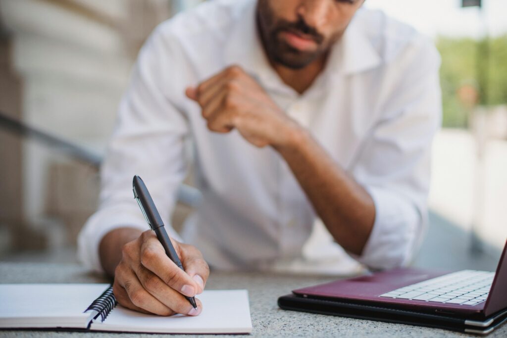 Man writing in a notebook next to a laptop, symbolizing the planning process behind a Content Writer Resume Template.