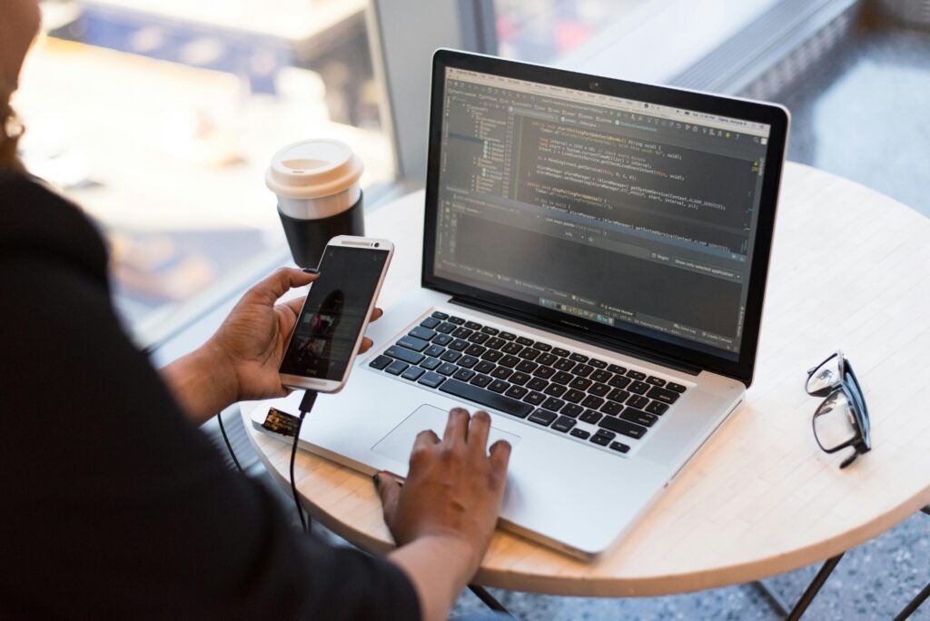 Person coding on a laptop while holding a smartphone, representing tech solutions that help manage outsourced payroll costs.