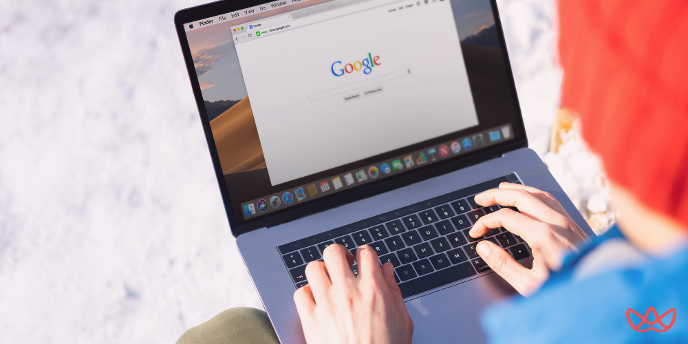 A person wearing a red beanie and a blue jacket typing on a laptop with the Google search page open on the screen, against a blurred white background suggesting snow or a bright outdoor setting. The laptop screen also shows the text "Outsource SEO A Complete Guide for 2025."