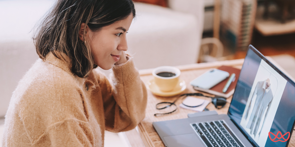 A woman with dark hair and a light brown sweater looking attentively at a laptop screen, with a cup of coffee, phone, and glasses on the desk. The laptop screen shows an image of clothing, and text reading "Outsourced Accounts Receivable The Complete Guide for 2025" is visible in the bottom left corner.