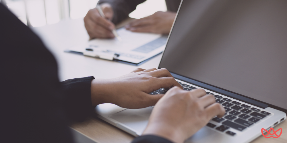 Close-up of a person typing on a laptop, with another person's hands and a document on a clipboard visible in the background, suggesting a business meeting or collaborative work session. The background text on the image reads "Payroll Services Fees & Outsourcing Costs Explained."