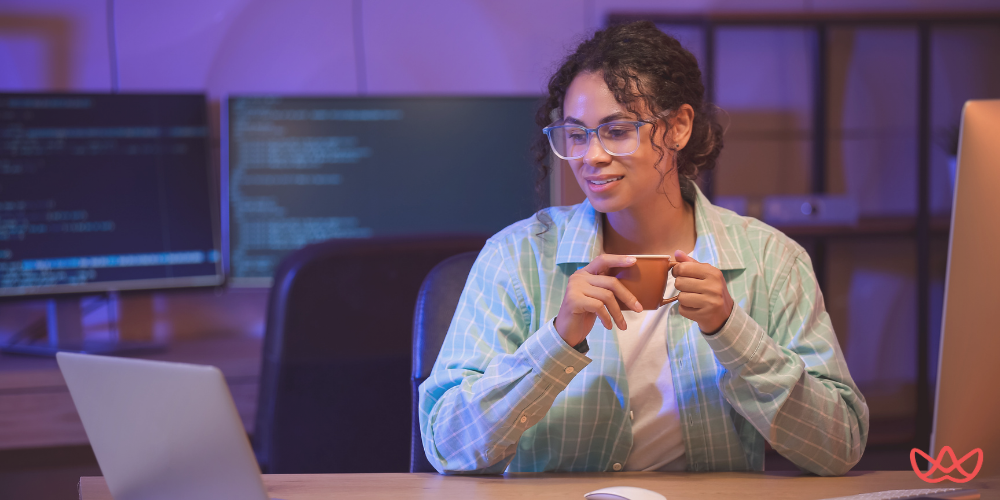 A smiling person with curly hair and glasses holding a cup while looking at a laptop in a dimly lit office with multiple computer monitors displaying code in the background. The image also contains the text "QA Outsourcing Services & Companies".