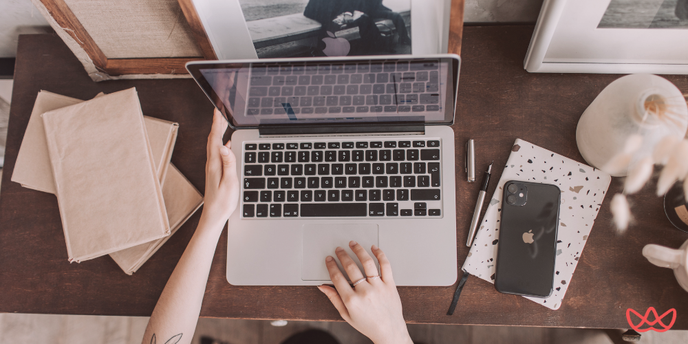 Overhead shot of a person's hands on a laptop keyboard and touchpad, with a smartphone, notebook, and stack of books on a desk, suggesting a workspace for managing finances or business operations. The laptop screen shows the text "Top Outsourced Accounting Services & Trends in 2025."