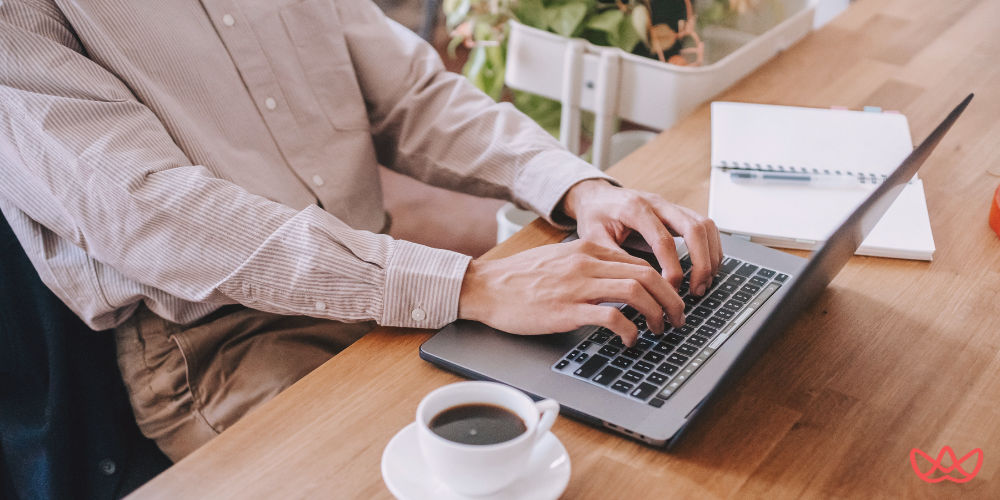 A person wearing a striped shirt and tan pants typing on a laptop at a wooden desk, with a cup of coffee and a notebook nearby, representing a professional working environment. The image also contains the text "What Is an Executive Assistant".