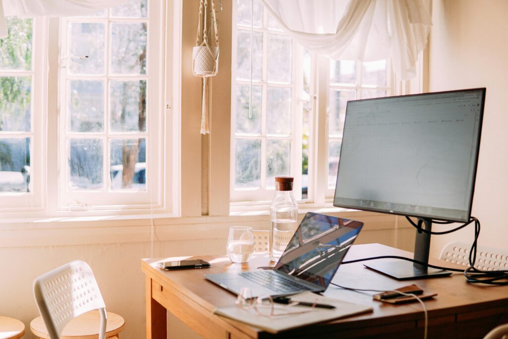 A well-lit home office setup with a laptop, an external monitor, a smartphone, and a water bottle on a wooden desk, positioned in front of large windows with sunlight streaming in, suggesting a remote work environment.