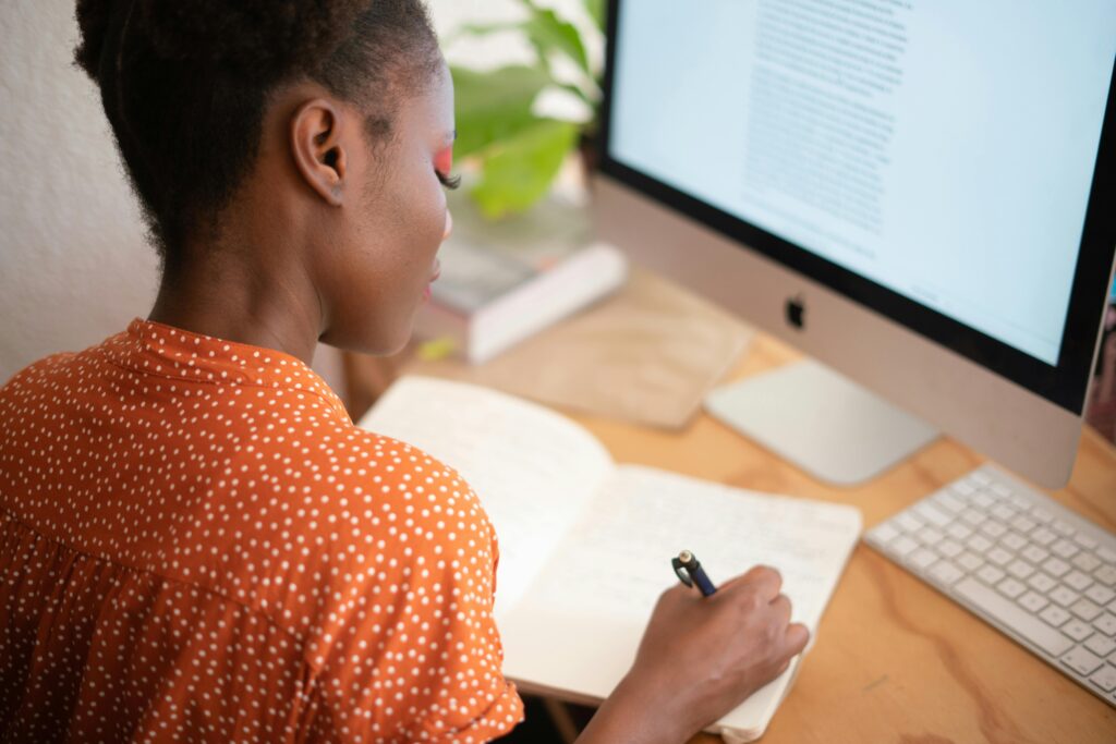 A woman with dark skin and an orange polka-dot shirt writing in a notebook while looking at a computer monitor showing text, suggesting a person working on a project, learning, or strategizing.