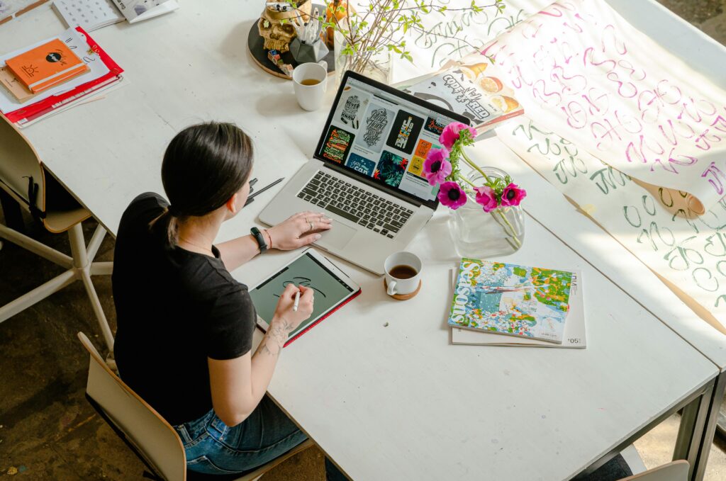 An overhead shot of a person working at a spacious desk, using both a laptop and a drawing tablet with a stylus, surrounded by creative materials, a vase of flowers, and a cup of coffee.