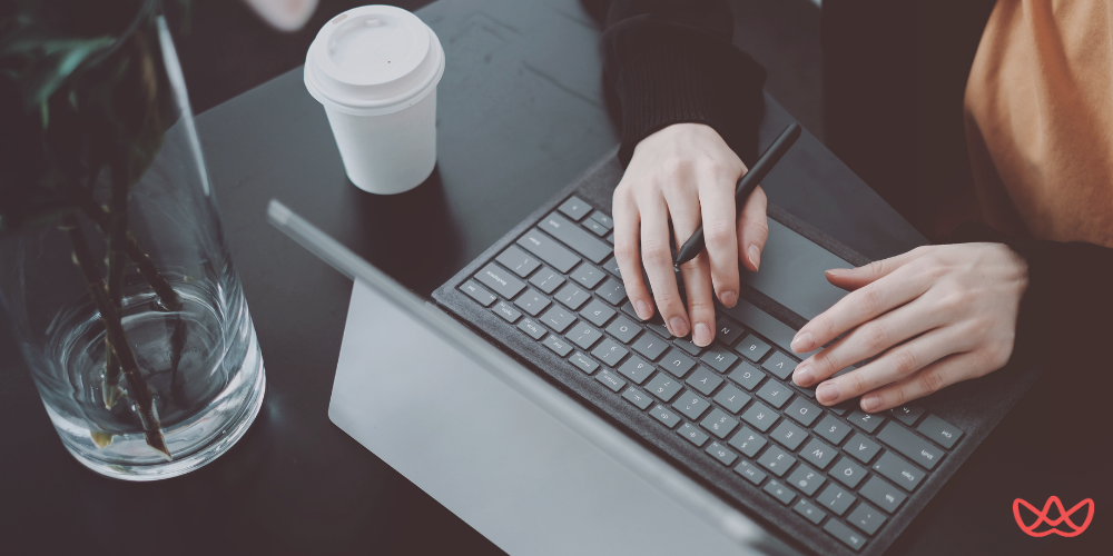 A close-up overhead shot of a person's hands using a stylus on a laptop's keyboard, with a white coffee cup and a glass vase with dark plant stems visible on a dark surface.
