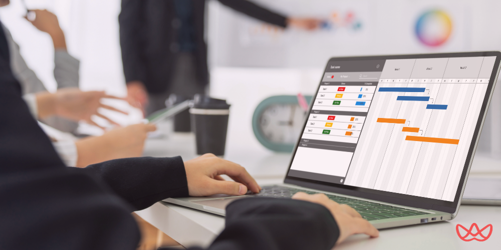 A laptop showing a Gantt chart or project timeline on its screen, with a group of people collaborating in the background at a meeting table.