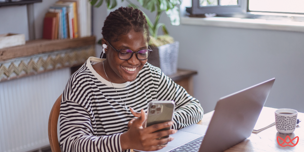 A woman with dark skin and braided hair, wearing eyeglasses and a striped long-sleeve shirt, smiling and looking at her smartphone while wearing earbuds, with a laptop and a coffee cup on a table in front of her. The image also contains the text "Remote Closing What It Is & How to Start a Career in 2025."