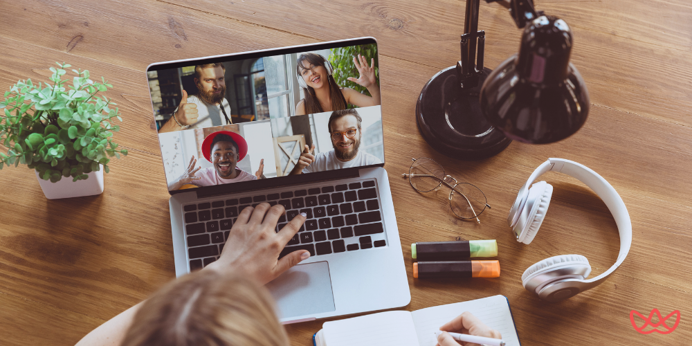 An overhead shot of a person's hands typing on a laptop during a video conference call showing multiple smiling faces, suggesting a remote team meeting or virtual interview. A plant, desk lamp, headphones, and highlighters are also visible on the wooden desk.