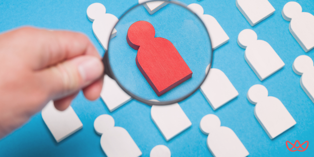 A magnifying glass held by a hand, focusing on a single red human-shaped figure among many white human-shaped figures on a blue background, symbolizing the process of selecting a specific individual from a group. The image also contains the text "Remote Staffing Agency Your Guide to Hiring Global Talent."