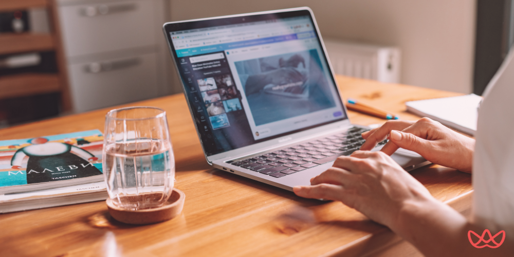 A close-up of a person's hands typing on a laptop keyboard, with a glass of water and books on a wooden desk, suggesting work in a home office or study environment. The laptop screen displays a creative design or content editing interface.